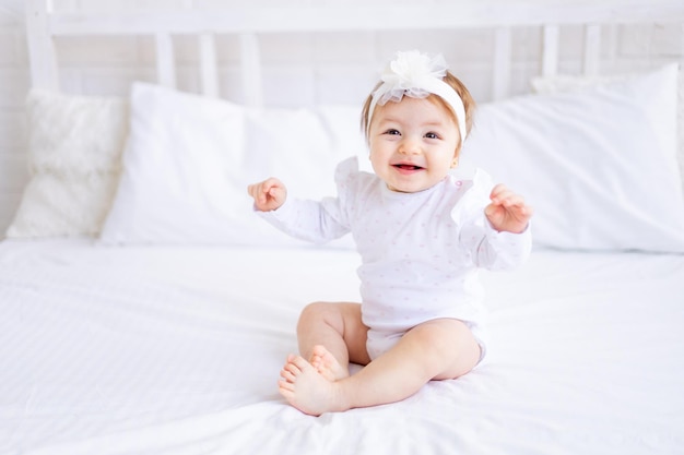 Cute baby girl sitting on the bed in white clothes and with a bow on her head funny little baby on a cotton bed at home and smiling baby products concept