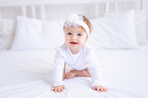 Cute baby girl sitting on the bed in white clothes and with a bow on her head funny little baby on a cotton bed at home and smiling baby products concept