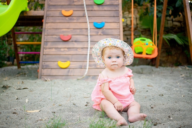 Cute baby girl sitting on the sand on the playground in summer in a panama hat and bare feet