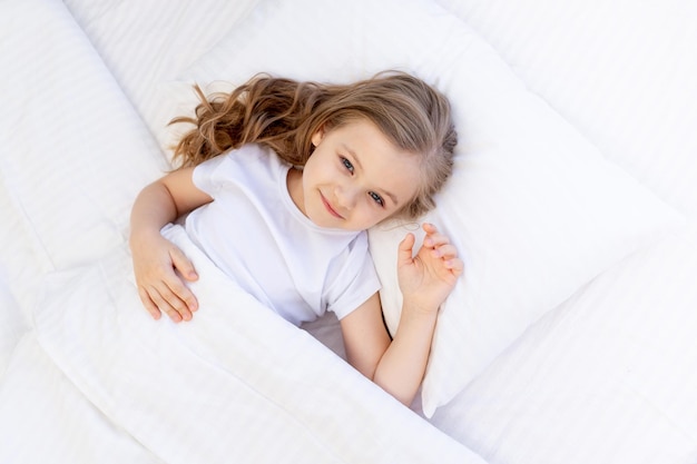 Милая девочка спит на кровати на белой хлопковой подушке под одеялом, здоровый детский сон ночью