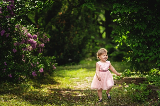 Cute baby girl on sunny day. Little girl with blond hair in pink dress smiling among blossoming trees.