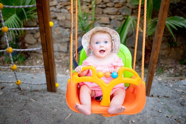 Cute baby girl swings on a swing and laughs in the summer in a panama hat