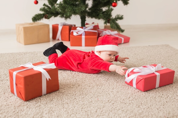 Cute baby girl wearing santa claus suit crawling on floor over Christmas tree. Holiday season.