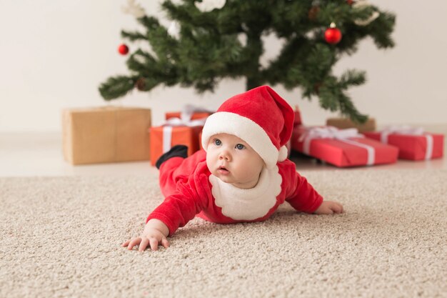 Cute baby girl wearing Santa Claus suit crawling on floor next to Christmas tree