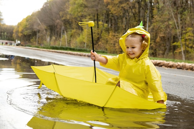 Cute baby girl wearing yellow stylish raincoat pink rubber boots sitting in umbrella in a puddle.