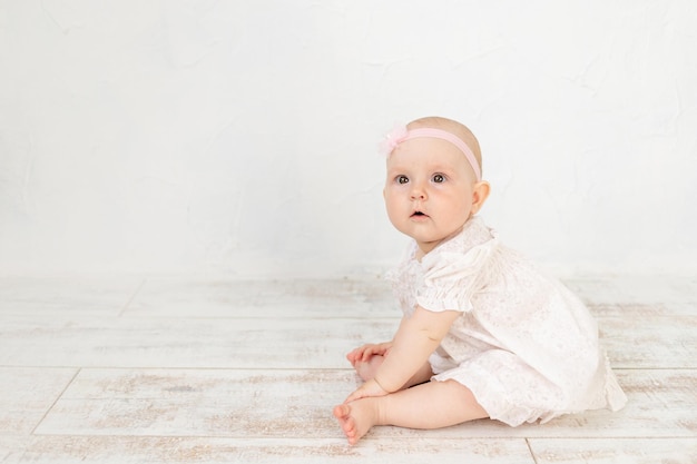 하얀 드레스를 입은 귀여운 여자 아이는 6개월 동안 바닥에 앉아 밝은 방과 텍스트를 위한 공간에 앉아 있습니다.