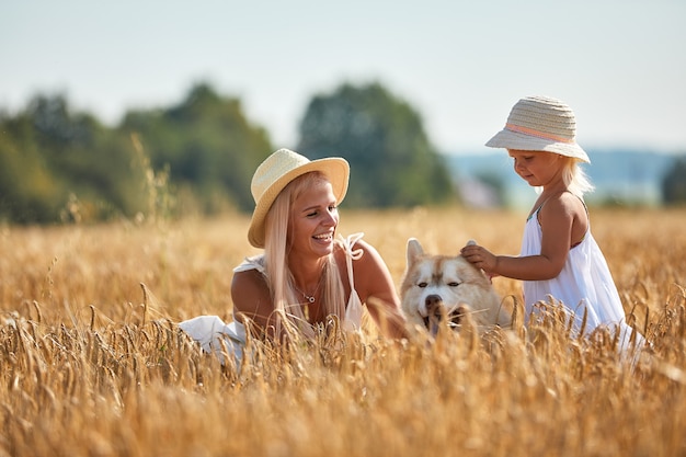 Cute baby girl with mom and dog on wheat field. Happy young family enjoy time together at the nature