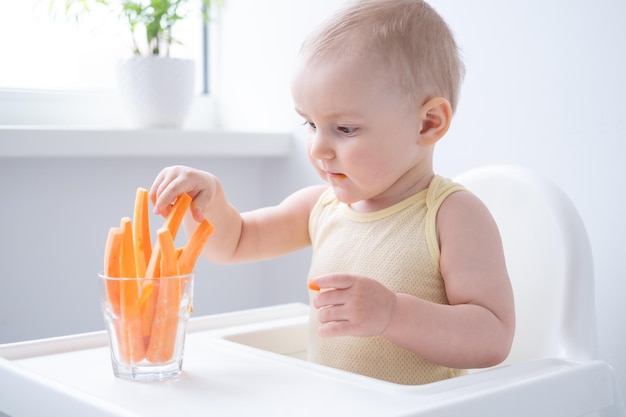 cute baby girl in yellow bodysuit sitting in childs chair eating carrot slices