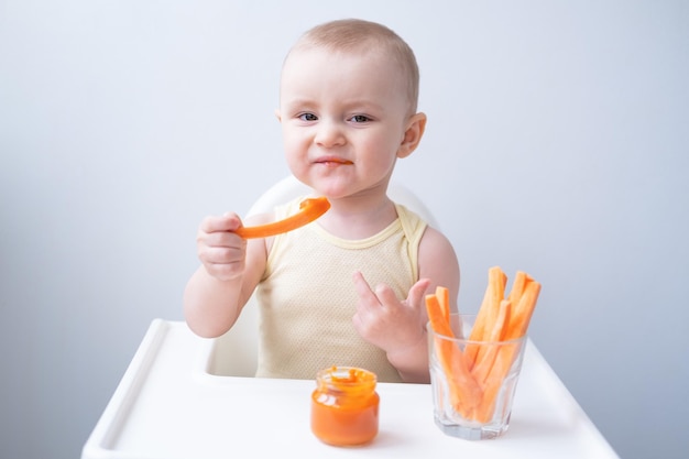 cute baby girl in yellow bodysuit sitting in childs chair eating carrot slices