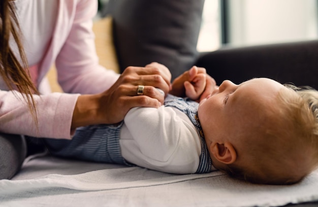 Cute baby and his mother holding hands while doing infant development exercises on the sofa