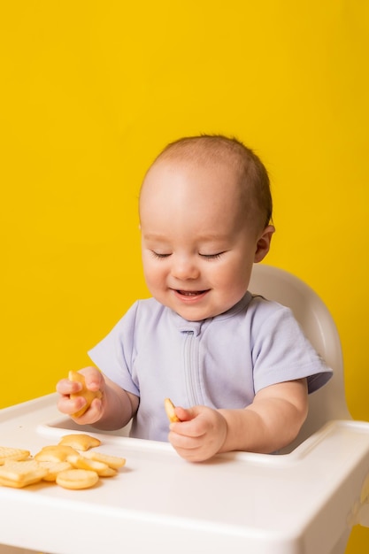 A cute baby is sitting in a child39s chair eating cookies Yellow background Baby food space for text