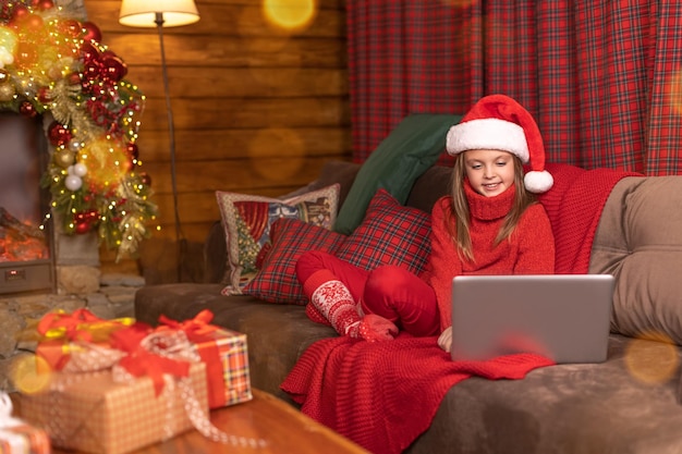 Bambina carina con un cappello da babbo natale seduta sul divano guardando un laptop con un sorriso prima di natale