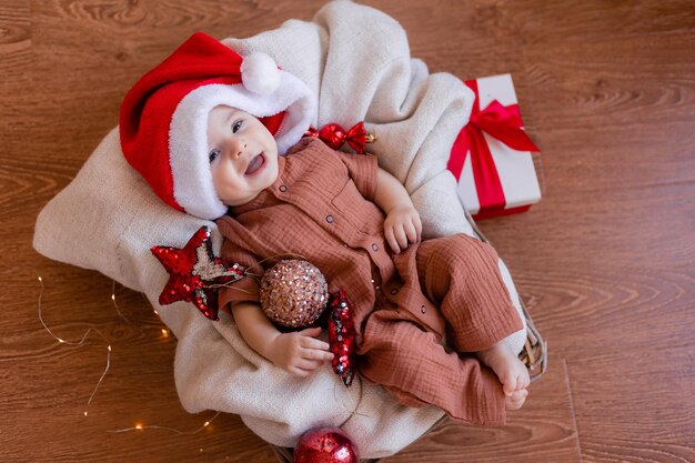 サンタの帽子をかぶったかわいい赤ちゃんが床の毛布に横たわって、彼の手にクリスマスボールを持っています。幸せな休日。冬、新年。高品質の写真