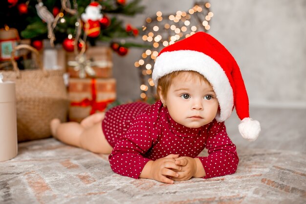 かわいい赤ちゃんサンタは贈り物を持ってクリスマスツリーの近くに家に座っています