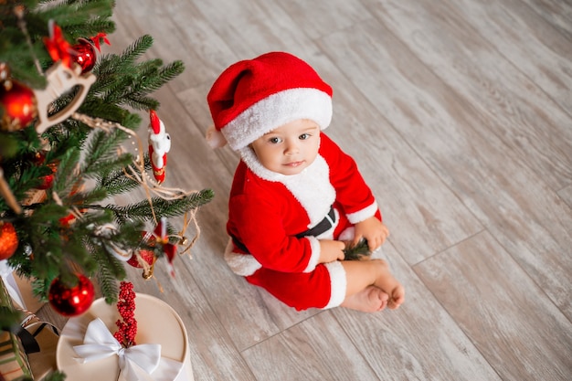 Cute baby Santa sits at home near the Christmas tree with gifts