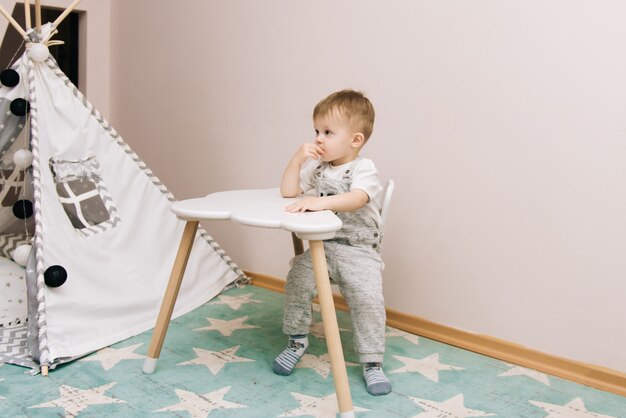 Photo cute baby sitting at the table and eating in the nursery white, gray and blue. near the teepee and a bag of toys