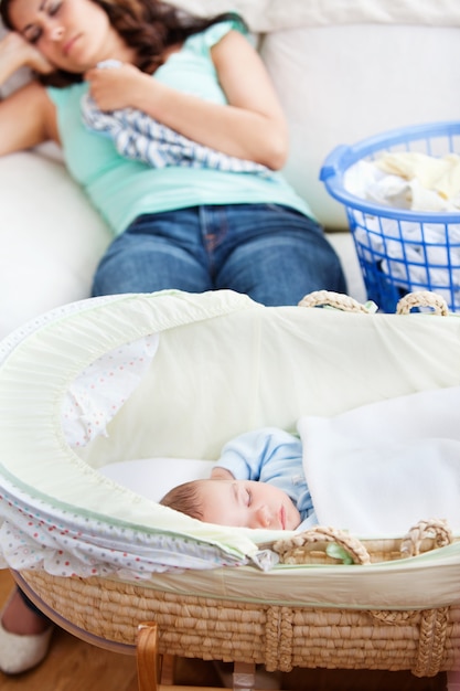 Cute baby sleeping in his cradle with his mother lying on the couch in the background