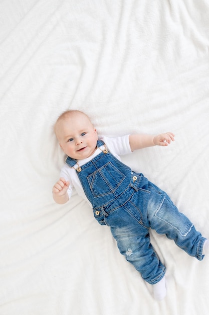 Cute baby smiles lying on a white bed at home in a denim suit