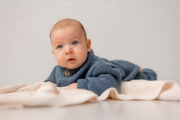 cute baby with blue eyes in blue knitted jumpsuit lies on his tummy on gray plaid solid background