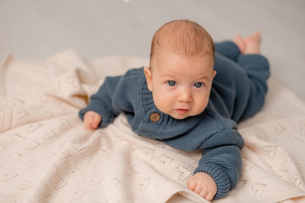 Cute baby with blue eyes in blue knitted jumpsuit lies on his
tummy on gray plaid solid background