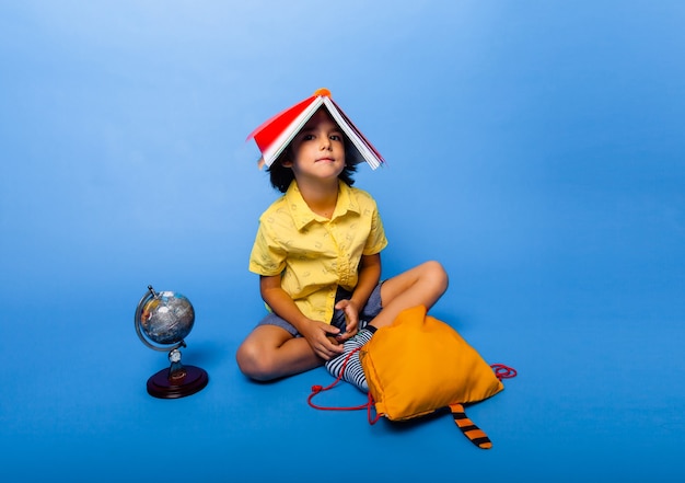 Foto bambino carino con un libro in testa. uno scolaro con un libro in testa si siede sul pavimento. preparazione per la scuola.