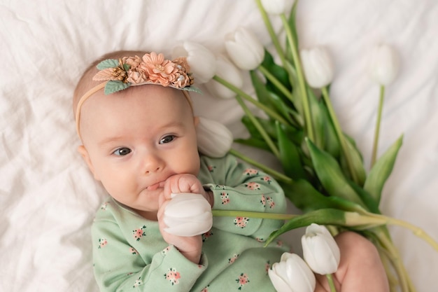 Cute baby with a floral headband and a green cotton bodysuit at home on a white bed with tulips. Spr