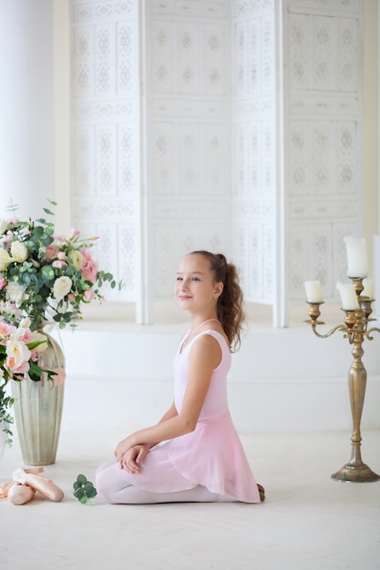 A cute ballerina in a ballet pink tutu and in points sits on the floor. Girl in the dance class.