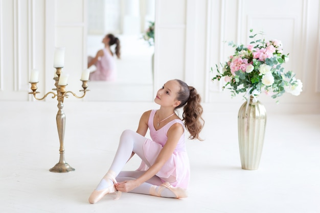 A cute  ballerina in a pink ballet costume is sitting on the floor and tying pointe shoes. Ballerina in the dance class.The girl is studying ballet. 