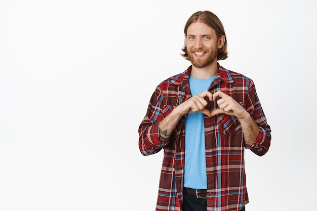 Cute bearded blond man smiling, showing heart I love you sign, like, express sympathy, standing in casual clothes on white