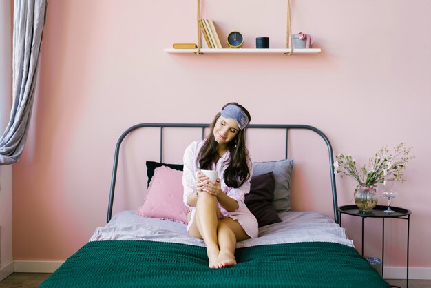 Cute beautiful brunette in a pink robe-shirt sits on the bed in the pink bedroom and holds a Cup of tea or coffee