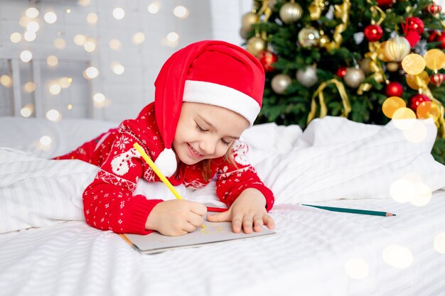 Cute beautiful girl child writes a letter to Santa Claus at the Christmas tree in a red sweater and hat on New Year's Eve or Christmas at home on a white bed