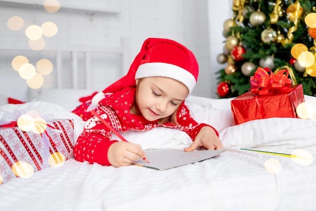 Cute beautiful girl child writes a letter to Santa Claus at the Christmas tree in a red sweater and hat on New Year's Eve or Christmas at home on a white bed