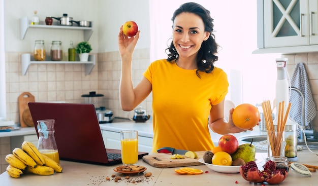 Cute beautiful and happy young brunette woman in the kitchen at home with fruits and laptop computer