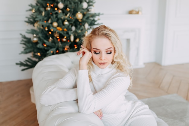 A cute beautiful happy young woman in a dress is resting while sitting on a white sofa near a Christmas tree