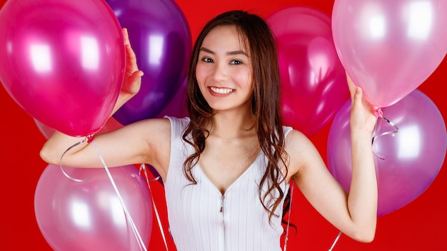 Carina e bella ragazza asiatica dai capelli lunghi scuri che tiene e gioca con mongolfiere colorate con un sorriso divertente e felice su sfondo rosso, riprese di luce in studio.