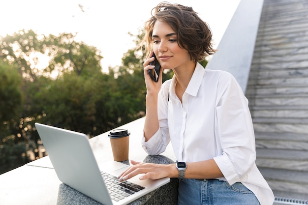 Cute beautiful woman standing outdoors using laptop computer talking by phone