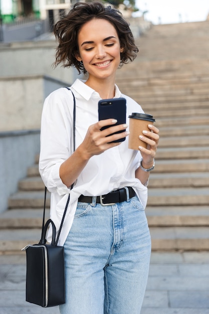 Милая красивая женщина гуляет по улице, используя мобильный телефон, держа кофе
