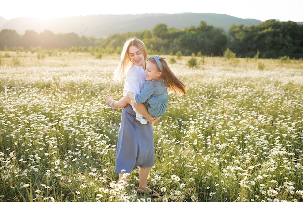 Cute beautiful young woman holding hands and play with little child girl walk in chamomile field with blooming flowers over nature background Family lifestyle concept Mom and baby daughter in grass