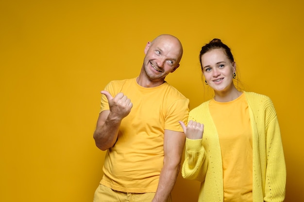 Cute benevolent man and woman in yellow clothes invite\
somewhere by pointing their thumb to the side