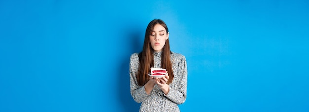 Cute birthday girl celebrating blowing candle on cake and making wish standing on blue background pa