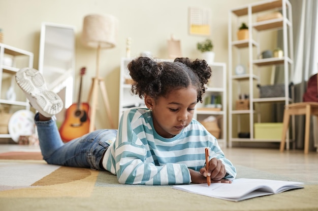 Photo cute black girl laying on floor at home and writing in notebook or diary