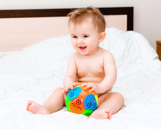 침실에 있는 침대에 앉아 공을 가지고 노는 귀여운 금발 아기. 공을 가지고 노는 6개월 된 행복한 아기