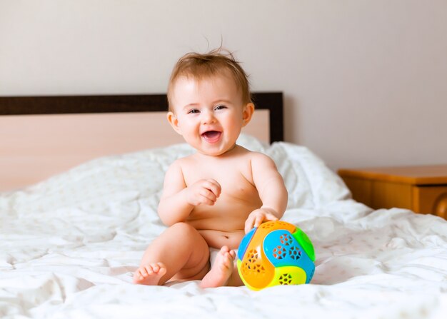 Cute blond baby playing with a ball sitting on the bed in the bedroom. happy baby 6 months old playing with a ball