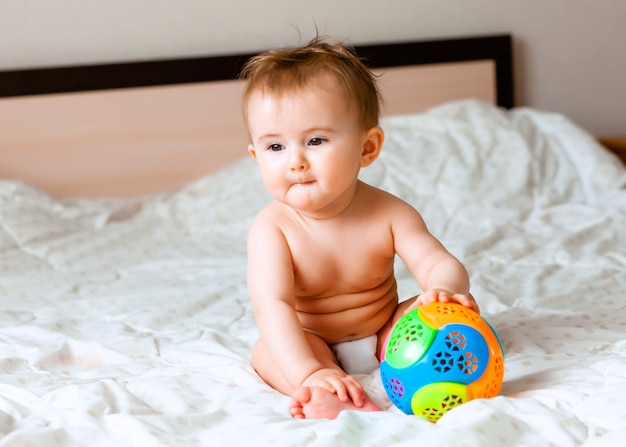 Cute blond baby playing with a ball sitting on the bed in the bedroom. happy baby 6 months old playing with a ball