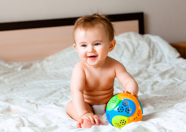 Cute blond baby playing with a ball sitting on the bed in the bedroom. happy baby 6 months old playing with a ball