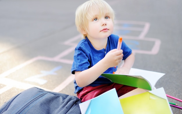 Cute blond boy doing homework sitting on school yard after school with bags laying near.