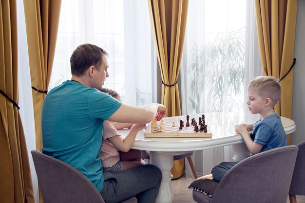 A cute blond boy and a girl play chess with their father have fun together at home A man teaches children to play a game play a board game play chess Family times