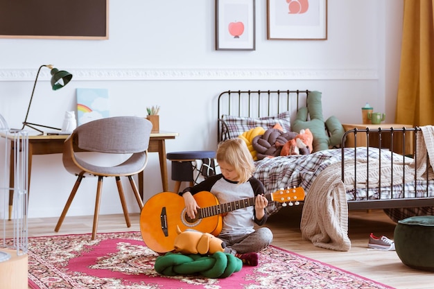 Милая блондинка сидит на полу в своей модной спальне и учится играть на гитаре.
