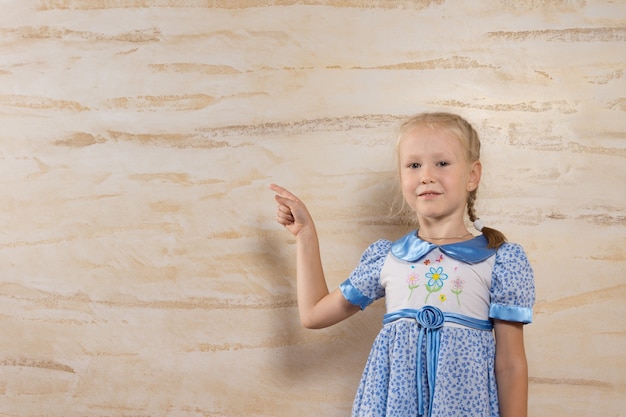 Cute blond girl wearing a dress with floral pattern wile pointing to the wooden wall behind her