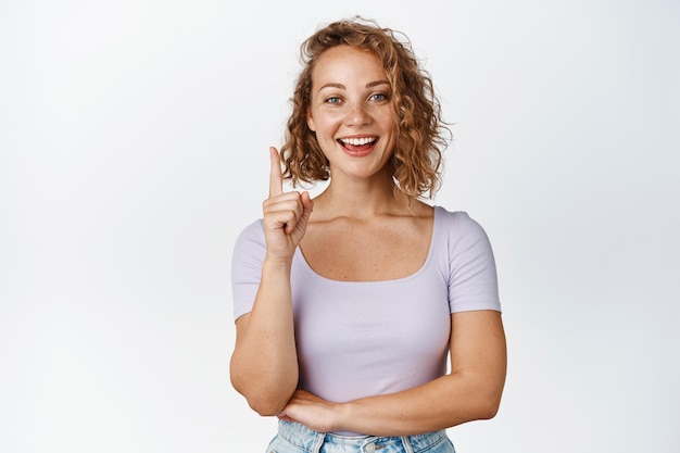 Cute blond girl with short haircut, pointing finger up and smiling, showing advertisement, standing in casual clothes over white background